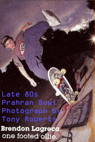 Photo taken in the early evening of Brendon Lagreca doing a big Ollie North at Prahran Bowl. 
    He is leaping high in the air with his front foot off the board. Brendon is wearing baggy blue cotton pants, white Nike high-tops and a baseball cap.
    A bright camera flash is going off in the background, as the sky changes color to purple before night.