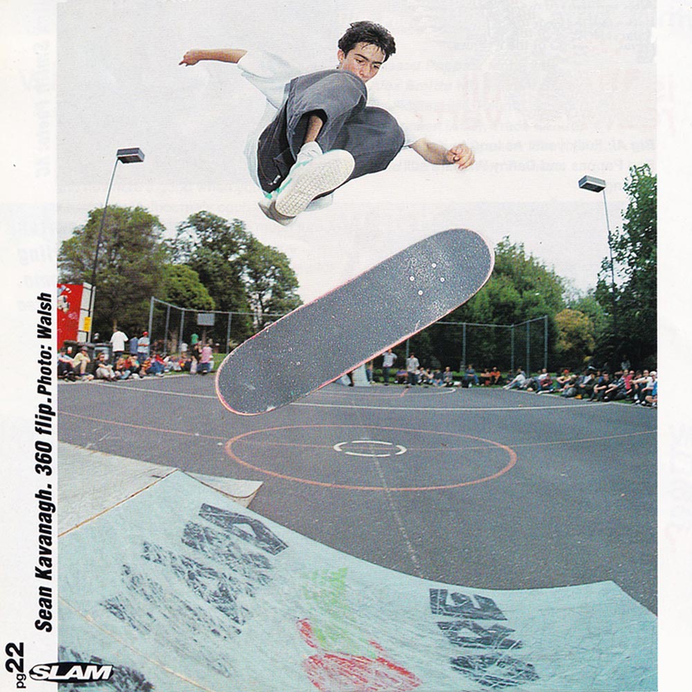 Photo of Sean Kavanagh doing a Tre Flip in the air above two ramps connected to a platform.
        He is wearing Baggy charcoal colored jeans with Light Green Puma Clydes. A crowd of onlookers watch in the background.
