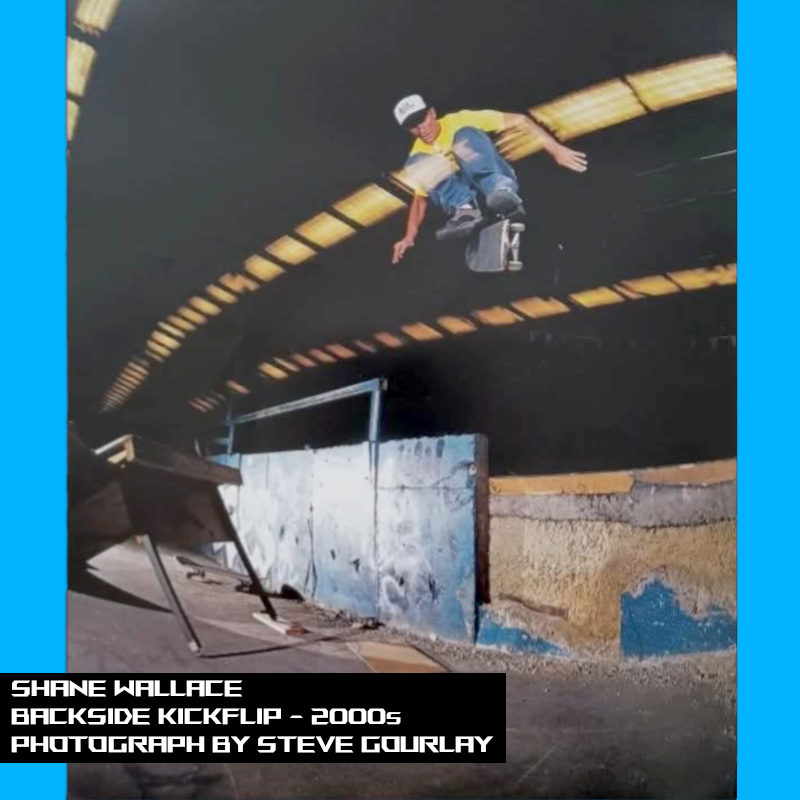 Shane Wallace doing a large backside kickflip off a table that has been made into a jump ramp.
  He is clearing a large gap onto a loading dock under the soft lights of an indoor warehouse.
